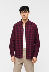 Νέος άνδρας που φοράει κόκκινο και μαύρο πουκάμισο με κουμπιά πάνω από λευκό μπλουζάκι και μπεζ παντελόνι, στέκεται μπροστά σε απλό φόντο.