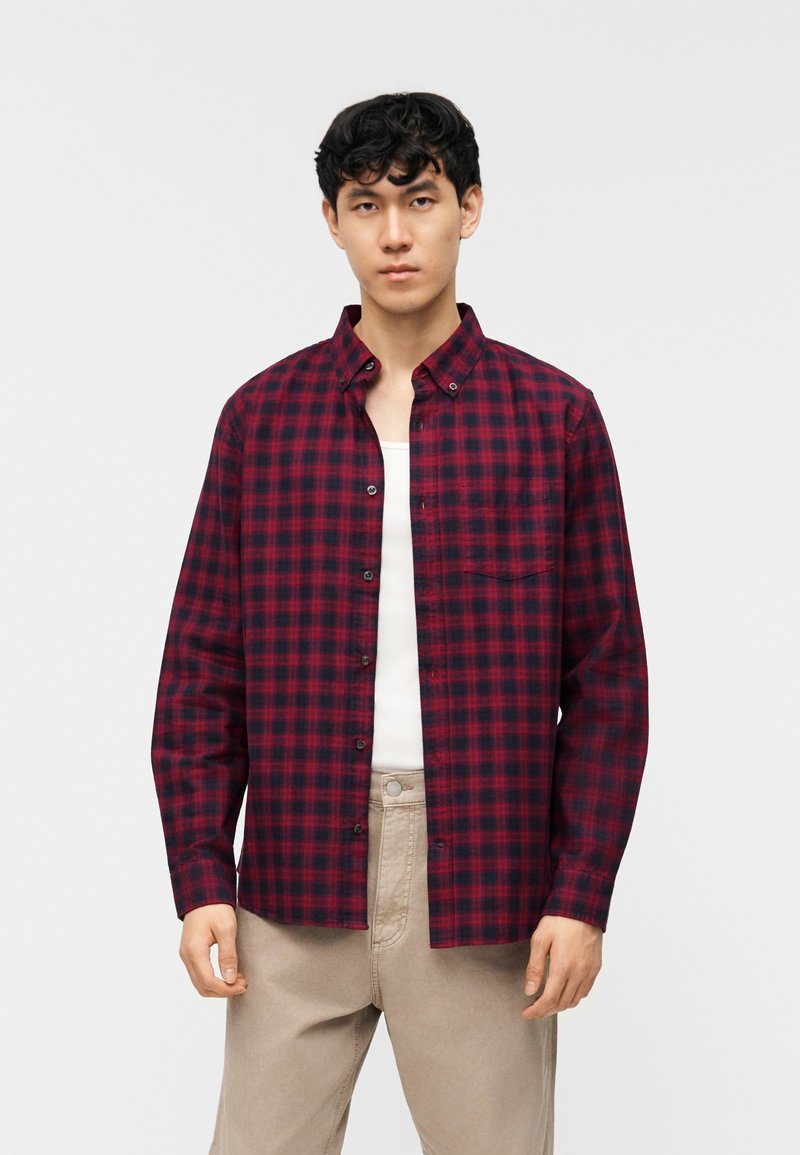 Νέος άνδρας που φοράει κόκκινο και μαύρο πουκάμισο με κουμπιά πάνω από λευκό μπλουζάκι και μπεζ παντελόνι, στέκεται μπροστά σε απλό φόντο.