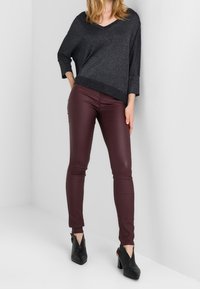 Czarny, błyszczący sweter z dekoltem w serek, zestawiony z bordowymi dopasowanymi spodniami. Spodnie mają gładką fakturę, a całość dopełniają czarne botki na obcasie.