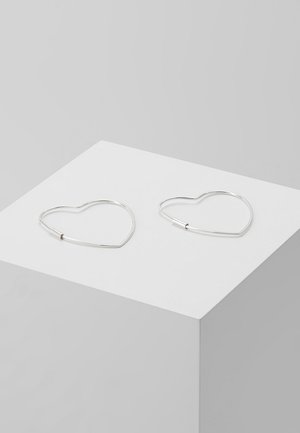 Deux boucles d'oreilles argentées en forme de cœur posées sur un socle blanc contre un fond gris clair.