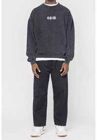 Osoba oblečená v tmavé oversized mikině se bílým japonským textem, černých kalhotách a bílých teniskách, stojící na jednobarevném pozadí.