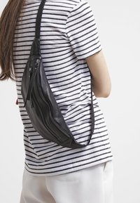 Persona que lleva una mochila de cuero negro cruzada en la espalda sobre una camisa de rayas blancas y negras y pantalones blancos.