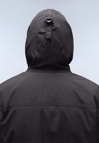 Personne portant une veste à capuche foncée avec une boucle à l'arrière de la capuche, vue de dos sur un fond clair uni.