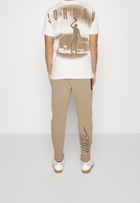 Vit t-shirt med bruna tryck, med en silhuett av en basketspelare och "JORDAN" text. Beiga sweatpants med svart "NIKE" logotyp. Vita skor.