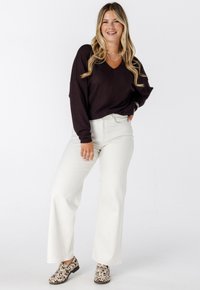 Femme souriante aux longs cheveux blonds, portant un pull en V marron foncé, un pantalon large blanc et des mocassins à motifs, se tenant avec une main sur la hanche.