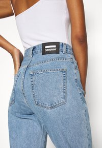Jeans de denim azul claro com uma cintura média, bolsos traseiros e um rótulo preto com "DRDENIM" na cintura. Textura desgastada.