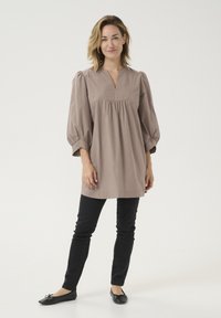 Tunic bej deschis, cu decolteu în V, mâneci bufante și detalii pliate. Asortată cu pantaloni negri subțiri și balerini negri. Țesătură din bumbac neted.