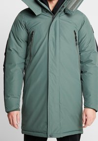 Grön isolerad parka med hög krage, dragkedja fram och två sidofickor med dragkedja. Har svarta detaljer på ärmar och mudd.