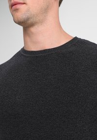 Dunkelgrauer Strickpullover mit rundem Halsausschnitt. Die Textur wirkt weich und leicht grob, mit einem schlichten, klassischen Design.