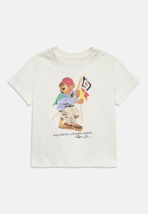 Biele detské tričko s gombíkovým zapínaním na ramene, s medveďom v červenej čiapke držiacim námorné vlajky, označené nápisom "Polo Bear by Ralph Lauren."