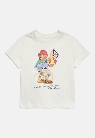 Λευκό παιδικό μπλουζάκι με κουμπωτό ώμο, που απεικονίζει μια αρκούδα με κόκκινο καπέλο να κρατά ναυτικές σημαίες, με την επιγραφή «Polo Bear by Ralph Lauren».