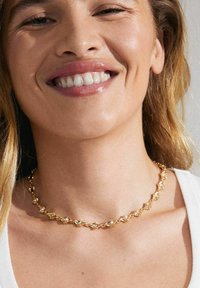 Gul choker halskæde med strukturerede led og små klare sten, prydet med et delikat, sammenflettet design. Båret om halsen.