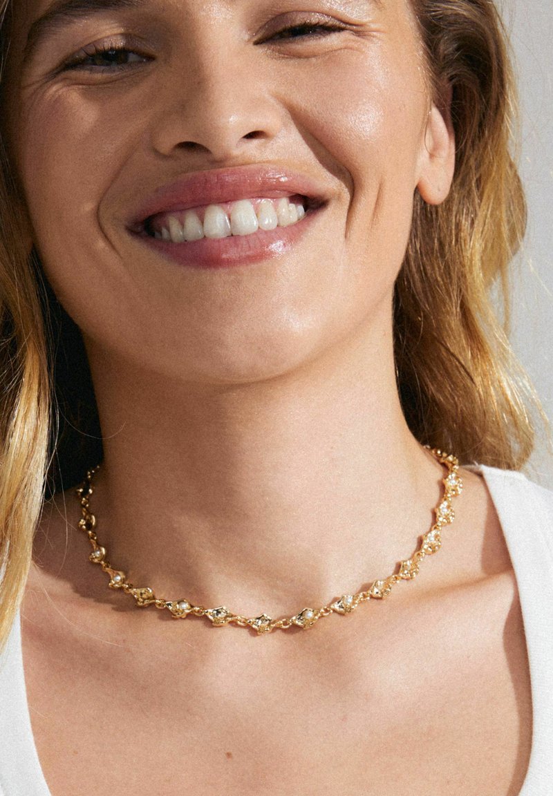 Gul choker halskæde med strukturerede led og små klare sten, prydet med et delikat, sammenflettet design. Båret om halsen.