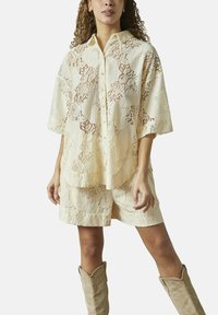 Creme blonder skjorte med knaplukning og korte ærmer, parret med matchende blonde shorts. Outfits fremhæver et struktureret blomstermønster.