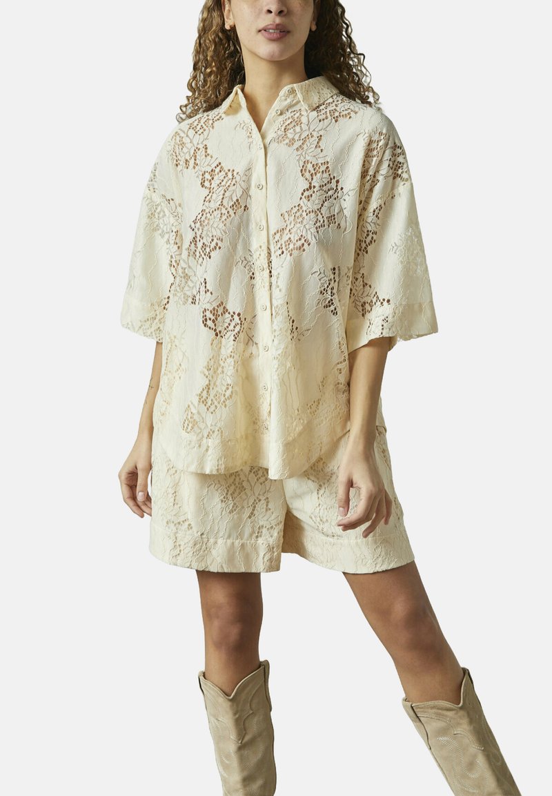 Creme blonder skjorte med knaplukning og korte ærmer, parret med matchende blonde shorts. Outfits fremhæver et struktureret blomstermønster.
