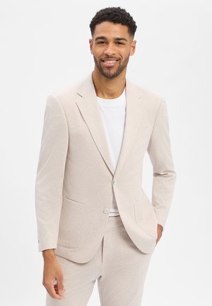 Man die lacht, gekleed in een lichtbeige colbert en bijpassende broek met een wit T-shirt, staand tegen een effen witte achtergrond.