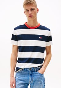 Stripete T-skjorte med marineblå og hvite horisontale striper, rød rund hals, liten logo-patch på brystet, kombinert med blå denimjeans.