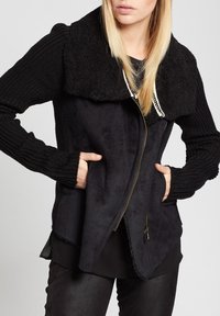 Femme portant une veste noire zippée avec un col texturé et des manches tricotées, mains dans les poches, sur un fond uni.