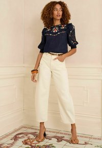 Navy embroidered blouse with puff sleeves, paired with loose cream trousers. The model wears brown heeled sandals and wooden bangles.