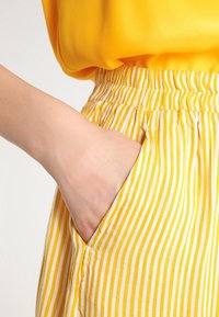 Tissu rayé jaune avec une ceinture élastique. La main repose dans une poche latérale. La texture semble lisse et légère.