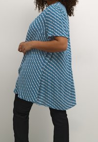 Blå bluse med mønster, der har et bølget hvidt design, korte ærmer, en rund kant og en afslappet pasform. Sammen med sorte bukser.