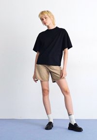 T-shirt nera oversize, pantaloni denim beige, calze bianche, scarpe nere. Il modello si staglia su uno sfondo semplice, mostrando uno stile casual e minimal.