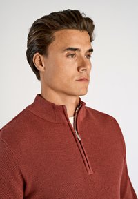 Punainen neulepuuvilla puoliveto, jossa on tekstuuri, varustettuna pystykauluksella ja harmaalla vetoketjydetaililla, käytetty valkoisen paidan päällä.