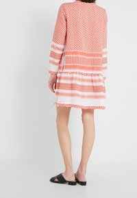 Vestido de patrones rojo y blanco con mangas largas, un dobladillo en capas y diseños geométricos variados; combinado con sandalias negras de deslizar.