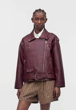 Vrouw met gevlochten haar, gekleed in een bordeauxrode leren jas over een wit shirt en bruine geruite korte broek, staand tegen een effen achtergrond.