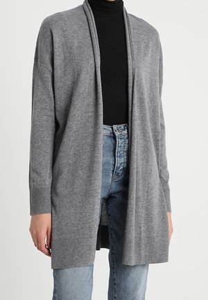 Person wearing a long, open-front gray cardigan over a black turtleneck and light blue high-waisted jeans.