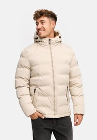 Veste rembourrée beige avec un col haut et une capuche r�églable. Présente des coutures horizontales, des poches zippées et une texture lisse.