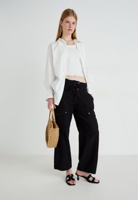 Vrouw poseert in een witte blouse over een witte cropped top, zwarte cargobroek, zwarte sandalen met hakken, terwijl ze een geweven handtas vasthoudt tegen een effen achtergrond.