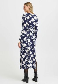Mörkblå klänning med vitt blommönster, långa ärmar och en rörlig design. Modellen har på sig svarta ankelskängor och genomskinliga svarta strumpbyxor.