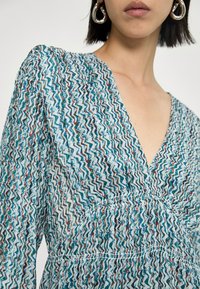 Kukallinen mekko, jossa on V-pääntie ja rypytetty yksityiskohta, sinisen, valkoisen ja punaisen sävyissä, kevyestä ja teksturoidusta kankaasta.