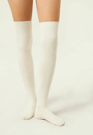 Chaussettes côtelées montantes en blanc cassé, fabriquées en matériau doux, avec un design texturé, un ajustement serré en haut et une zone de pied lisse.