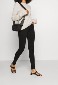 Suéter de punto beige claro, jeans ajustados negros, bolso negro de bandolera y sandalias de tacón con tiras negras. Textura sencilla y diseño ajustado.