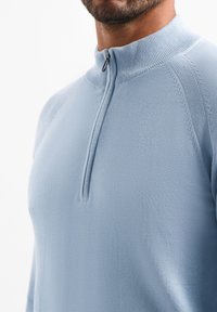 Lichtblauwe pullover met een ritssluiting en een opstaande kraag. Heeft een gestructureerd breipatroon en ribgebreide accenten bij de kraag en schoudernaden.