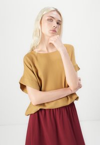 Sennepsgul kortermet bluse med bred utringning, kombinert med en luftig, burgunder skjørt. Glatt stoff med løs passform og elastisk midje.