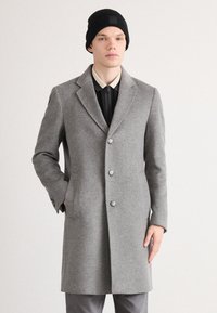 Cappotto grigio in misto lana con colletto a rovescio, cinque bottoni e tasche laterali. Indossato sopra una camicia nera e completato da un berretto nero.