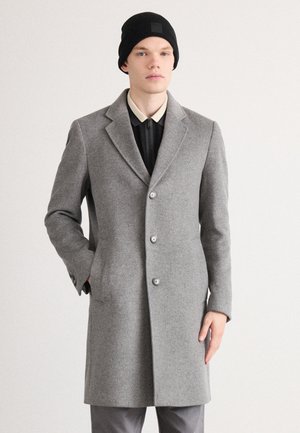 Manteau en mélange de laine gris avec un col cranté, cinq boutons et poches latérales. Porté sur une chemise noire et complété par un bonnet noir.