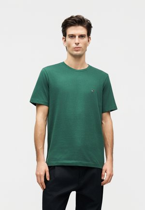 Νεαρός άνδρας στέκεται, φοράει ένα σκουροπράσινο κοντομάνικο μπλουζάκι και μαύρο παντελόνι μπροστά σε απλό ανοιχτό φόντο.