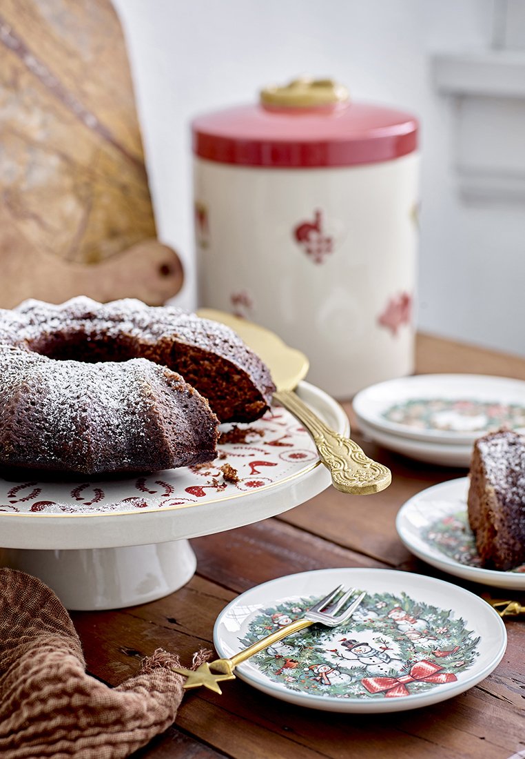Schokoladen-Bundt-Kuchen, bestäubt mit Puderzucker, auf einem gemusterten Teller, mit dekorativen Tellern und Gabeln auf einem Holztisch.