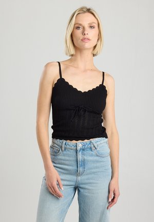 Black ribbed tank top with scalloped neckline and adjustable straps, featuring a front tie detail. Paired with light blue high-waisted jeans.