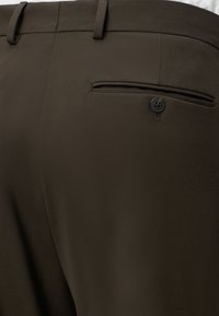 Gros plan d’un pantalon habillé marron foncé montrant la taille, les passants de ceinture et une poche arrière avec un bouton noir.