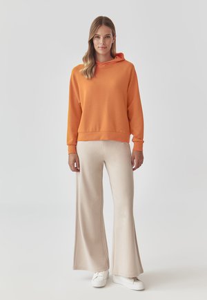 Felpa arancione con cappuccio, vestibilità morbida, polsini a costine, abbinata a pantaloni larghi color crema e sneaker bianche. Tessuto liscio, design informale.
