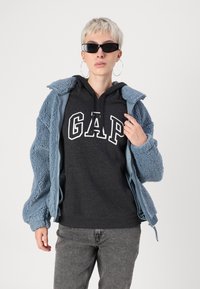 Blå sherpa-jakke med glidelås, brukt over en grå hettegenser med hvit "GAP" logo, sammen med grå jeans og svarte solbriller.