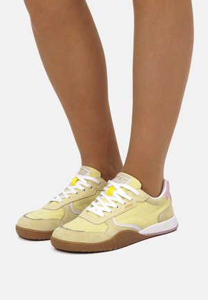 Benen met lichtgele sneakers met witte veters, bruine zolen en een vleugje roze op de hiel, staand op een wit oppervlak.
