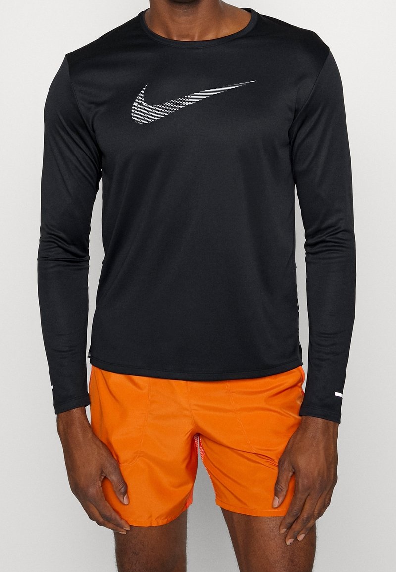 Mann trägt ein schwarzes langärmliges Nike-Shirt mit weißem Logo und leuchtend orangefarbenen Sportshorts, steht vor einem schlichten, hellen Hintergrund.