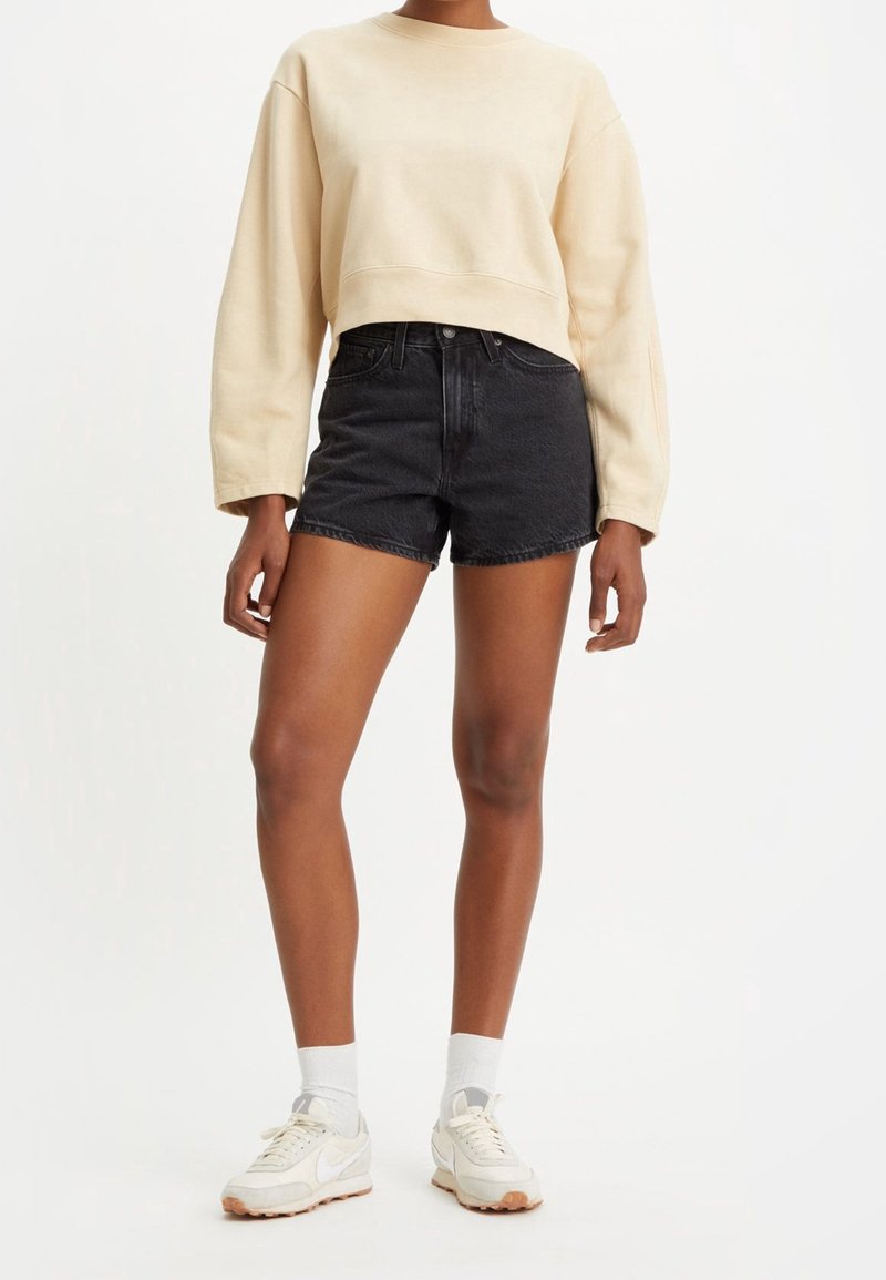 Personne portant un sweat-shirt court beige à manches longues, un short en jean noir taille haute, des chaussettes blanches et des baskets blanches, debout sur un fond blanc.