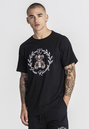 Młody mężczyzna z krótkimi włosami i tatuażami nosi czarną koszulkę z motywem misia w kratę i napisem "Gianni Kavanagh".